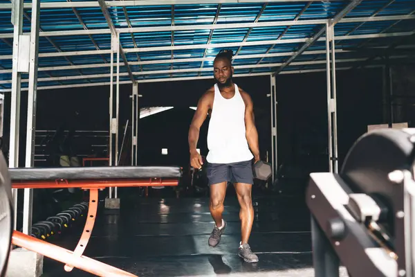 Full body of confident bearded African American male athlete in sportswear walking with dumbbell during workout in modern gym with equipment under dark metallic shade shelter