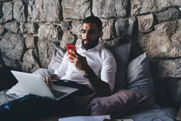 Serious bearded male in formal apparel sitting on bed with cushions and browsing mobile phone while working online on laptop with marketing plan