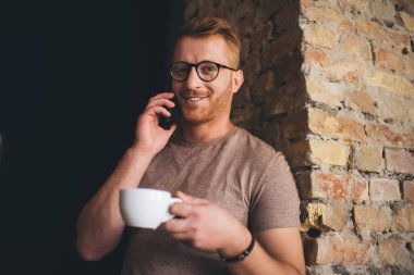 Kahve molası sırasında cep telefonu teknolojisiyle iletişim kurarken kameraya gülümseyen, cep telefonuyla konuşan, optik gözlüklü mutlu sakallı bir adamın yarım boy portresi.