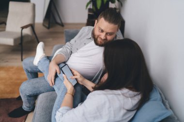 Yüksek açılı, sevecen genç bir erkek kucağında tanınmayan kadın bacaklarına dokunurken rahat bir kanepede dinleniyor ve kız arkadaşı evde parmaklarıyla cep telefonunu karıştırıyor.
