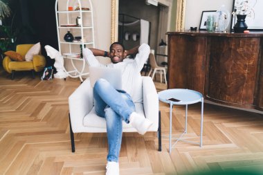 Yukarıdan, akıllı telefonuyla kanepenin yanındaki koltukta dinlenen ve dizüstü bilgisayara bakıp oturma odasında komik videolar seyreden neşeli Afrikalı Amerikalı bir erkek.