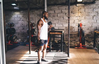 Spor kıyafetli güçlü Afrikalı Amerikalı sakallı bir adamın iki eliyle dambılları kaldırırken ve spor salonunda duvar duvarı ile çalışırken tüm gövdesinden görünüşü.