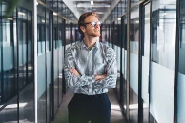 Resmi giysiler ve gözlüklerle gülümseyen genç sakallı erkek çalışan modern koridorda kolları çaprazlanmış, cam kapılar diğer tarafa bakıyor.