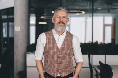 Kendine güveni tam, şık giyinmiş olgun bir erkek. Ofiste elleri ceplerinde, iç mimarisi bulanık bir şekilde kameraya bakıyor.