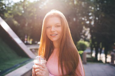 Güneşli bir günde elinde bir şişe limonata ile parkta dinlenirken gülümseyen ve kameraya bakan genç bir kadın sokakta bulanık yeşil ağaçların yanında.