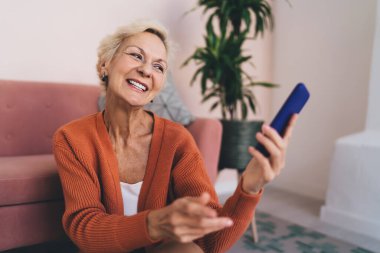 Keyifli, dişlek gülüşü olan, rahat oturma odasında otururken akıllı telefon kullanarak selfie çeken neşeli, yaşlı bir bayan.