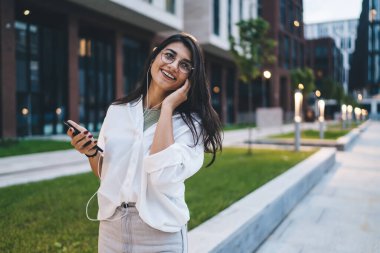 Sokakta akıllı telefonlarda kulaklıkla müzik dinlerken dişlek gülüşlü gözlüklü, sıradan giyinmiş genç bir kadın.