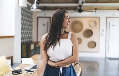 Kafe 'de çalışırken kahve fincanı akıllı telefonu ve laptopuyla masada durup gülümseyen ve yüzünü çeviren gündelik kıyafetlerle mutlu, genç, etnik, serbest bir bayan.