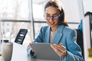 Modern ofiste parlak bulanık bir arka plana karşı proje üzerinde çalışırken bir fincan kahve ve tabletle masada oturan resmi kıyafetli ve gözlüklü pozitif kadın serbest yazar.
