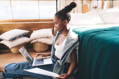 Afro-Amerikalı kadın, saçı örgülü, yatağın yanında oturuyor ve evde netbook 'u tarıyor.