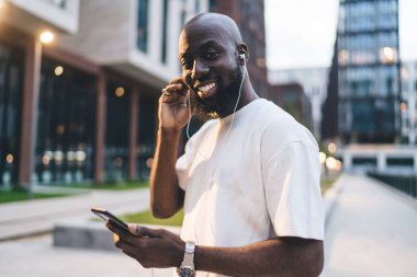 Pozitif genç, sakallı, beyaz tişörtlü Afro-Amerikan erkek. Gülümsüyor ve kameraya bakıyor. Cep telefonuyla sörf yapıyor ve sokakta kulaklıkla müzik dinliyor.