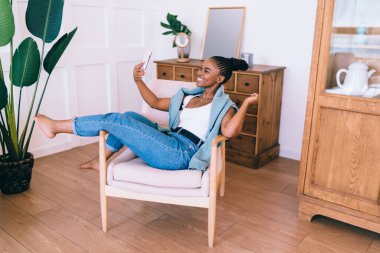Koltuğunda dinlenirken gülümseyen ve cep telefonundan selfie çeken çıplak ayaklı, Afro-Amerikan bir kadının tüm vücudu.