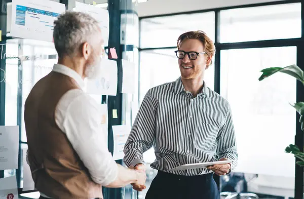 Positive redhead male employee shaking hands with aged colleague while standing near glass wall with business strategy after brainstorming project