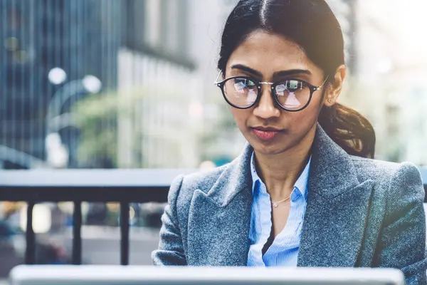 Close up of concentrated Indian female in eye wear for vision correction working on touch pad while sitting outdoors in downtown, Female entrepreneur reading news in network. Millennial business work