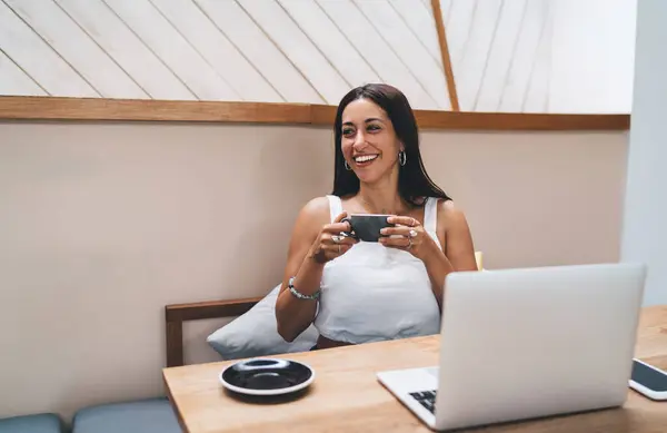 Smiling female freelancer wearing casual clothes having cup of tea looking away while working online with laptop in modern coffee shop