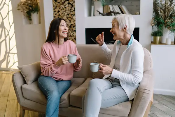 Positive diverse elderly women with mugs of hot drink smiling and looking at each other while having fun together during conversation