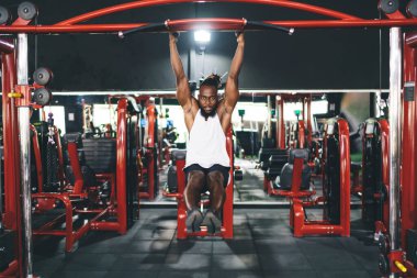 Spor giyim kuşamında genç, güçlü, sakallı Afrikalı Amerikalı erkek sporcu. Modern spor salonunda egzersiz yaparken esnek vücutlarıyla barfiks çekiyor.