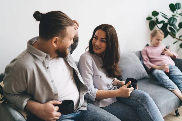 stock image Positive woman and man in casual outfits sitting on couch having cups of hot beverage communicating while daughter browsing cellphone