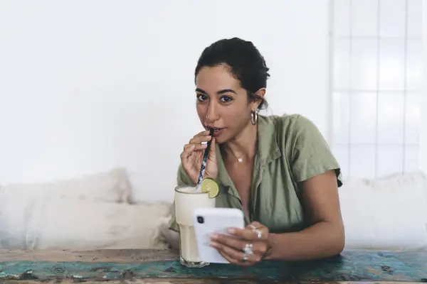 Cheerful young Hispanic female sitting at table and drinking cool milkshake with lime slice from glass while looking at camera and browsing smartphone