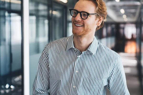 Positive crop young redhead male manager in striped shirt and glasses smiling while walking in modern office hallway during workday against blurred background
