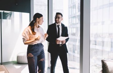Gözlüklü gülümseyen genç bir adam gazete tutuyor ve iş kurma ve ofiste yürüme hakkında bilgi paylaşırken bayan meslektaşına bakıyor.