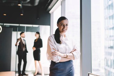 Resmi giysiler içinde başarılı bir bayan girişimci ofiste pencerenin yanında dururken çapraz kollarla kameraya bakıyor ve gülümsüyor.