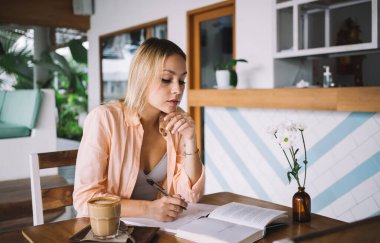 Elleri çenesine değen düşünceli genç bir bayan bir bardak kahveyle ahşap masada oturuyor ve defterine defterden notlar yazarken ve gün içinde kafede dinlenirken aşağıya bakıyor.