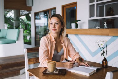 Gülümseyen genç bayan bir bardak kahveyle masada oturuyor ve kafeteryada boş zamanlarında gözlerini kaçırırken elindeki kalemle not alıyor.
