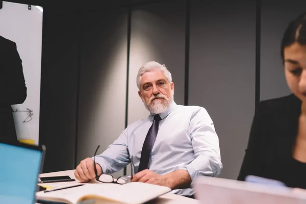 Self assured senior male entrepreneur in formal wear with gray hair and beard looking at camera while sitting at table during business meeting in boardroom in daytime