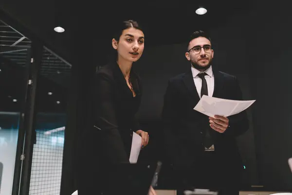 Low angle of confident office employees in formal dress standing with documents in hands near table and looking away while discussing on business presentation in office room