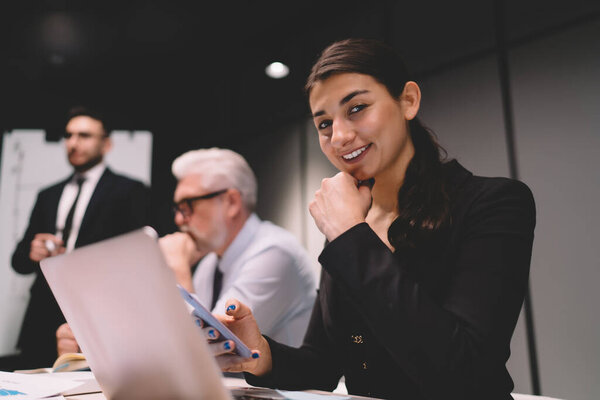 Позитивная молодая деловая женщина в формальной одежде сидит за столом со смартфоном и ноутбуком во время работы с коллегами в современном офисе
