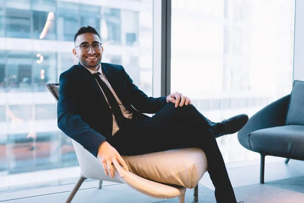 Portrait of friendly entrepreneur in classy clothes looking at camera and smiling while resting in comfortable armchair from work in spacious office