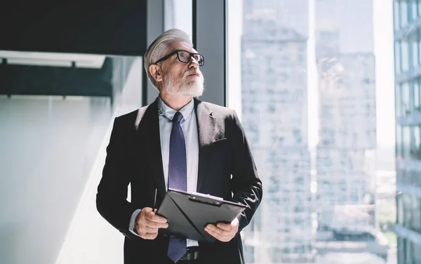 Confident aged businessman in formal clothes and eyeglasses standing near glass wall with sun reflecting clipboard and pen in hand while looking away at buildings outside in modern office