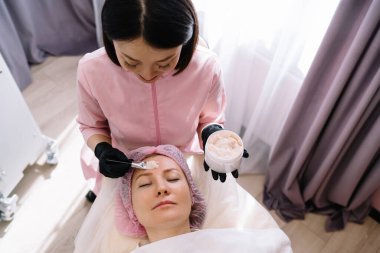 Asyalı güzellik uzmanı yüzüne fırçayla krem sürerken kapalı gözlerle masanın üzerinde uzanan kadının tepesinden.