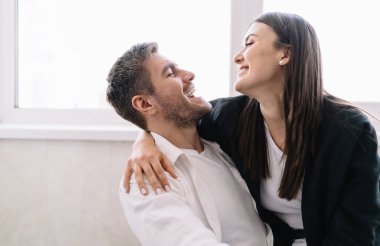 Çiftler birbirlerine bakıp gülümserken ve kucaklaşırken ve pencerenin yanında gündüz vakti parlak güneş ışığıyla ayakta dururken.