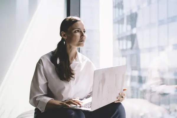 Pensive female executive in formal clothes sitting on windowsill and looking away thoughtfully while browsing laptop during work at modern office