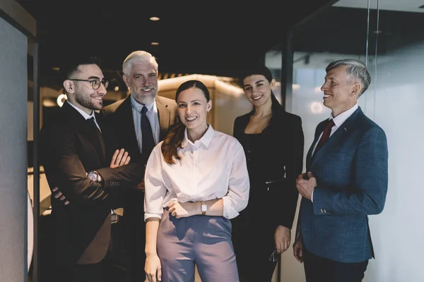 Positive smiling coworkers in elegant clothes standing near glass wall in modern workspace together while working in contemporary business center