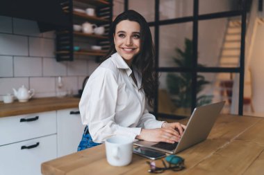 Modern mutfaktaki masada oturan beyaz gömlekli serbest çalışan genç, pozitif bir bayan internet sayfası taraması yapıyor.