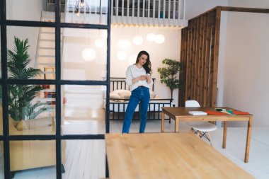 Yurt odasındaki ekrana bakarken, kitap ve cep telefonuyla masanın yanında duran, günlük kıyafetli genç kız öğrencilerden oluşan bir vücut.