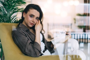 Kameraya bakarken koltukta kitap okuyarak takılan, şık, sıradan giysiler içinde genç esmerden oluşan camdan bir duvara bak.