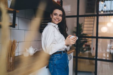 Günümüz mutfağında, cam duvarın yanında gülümseyen ve yüzünü çeviren bir bardak sıcak içecekle duran şık giysili pozitif kadın.