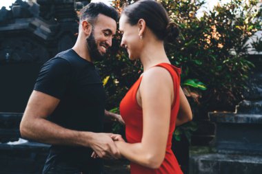 Side view of young man and woman looking at each other and smiling while standing near green bushes during romantic dance in garden clipart