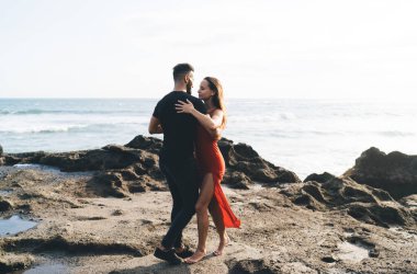 Kırmızı elbiseli genç adam ve kadının tüm vücut görüntüsü ve günlük giysiler. Parlak gün ışığında kayalık sahilde durup birbirlerine sarılıyorlar.