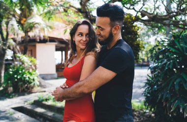 Side view of smiling young couple embracing while standing in green park and looking at each other near trees and bushes clipart