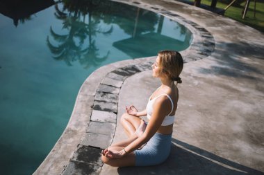 Chin Mudra 'da yüzme havuzunun kenarında oturan ve güneş ışığında lotus pozu veren ve yazın tropikal iklimin tadını çıkaran genç dişinin yan görüntüsü.