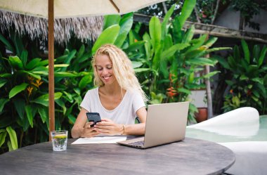 Gülümseyen genç bayan blogcu havuz başında limon parçalı dizüstü bilgisayarlı bir bardak suyla oturuyor ve gündüz vakti blog yazarken akıllı telefon ekranına bakıyor.