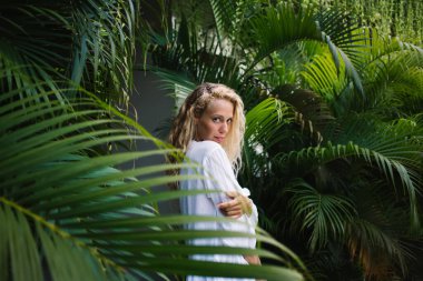 Beyaz elbiseli genç bir kadın, tropikal bahçede yeşil egzotik bitkilerin yanında kameraya bakarken.