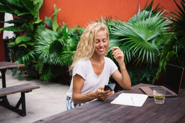 Günlük giysiler içinde neşeli genç bir bayan kafeteryada akıllı telefondan mesajlaşırken dizüstü bilgisayarı ve defteriyle ahşap masada oturuyor.