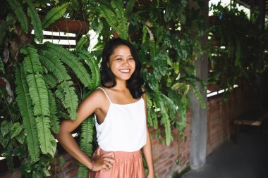 Olumlu etnik kadın modelin portresi kameraya bakıyor ve gülümsüyor yeşil egzotik bitkilerle tuğla duvara yaslanırken.