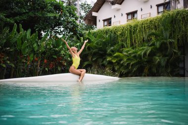 Mayo giymiş sıska genç bir kadın havuz kenarında kollarını kaldırmış oturuyor ve yaz tatilinin keyfini çıkarıyor.